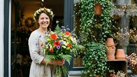 Young woman with curly blonde hair and fair complexion, wearing a light cardigan, holding a vibrant bouquet of red and orange flowers outside a flower shop with ivy-covered storefront. Use for florist marketing, small business promotion, or lifestyle content.