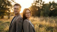 Professional couple in glasses standing back to back outdoors during golden hour sunset, smiling confidently in business attire for corporate teamwork concepts.