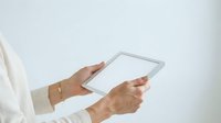 Woman with medium skin tone holding blank tablet device with both hands, wearing white long-sleeve shirt and gold bracelet. Suitable for technology, business, and digital communication concepts.
