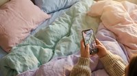 Person with medium skin tone holding smartphone taking photo of colorful bedding in pastel pink, green, and purple tones, overhead flat lay composition for content creation and social media.