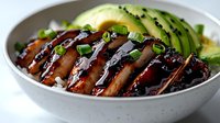 Sliced teriyaki chicken breast with dark sweet soy glaze, garnished with green onions and fresh avocado in white ceramic bowl, close up view of Asian cuisine.