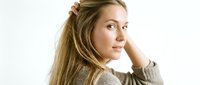 Young woman with long brown hair and fair complexion, wearing casual gray sweater, posing thoughtfully with hand near head against white background for portrait and lifestyle use.