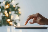Female hand touching digital tablet screen with blurred festive bokeh lights and green plant decoration in background, creating cozy atmosphere.