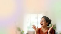 Woman with dark hair and medium skin tone wearing rust-colored top, smiling with hands raised in joyful gesture near bright window with natural light and blurred interior background.