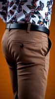 Close-up view of tan formal trousers with black leather belt and floral pattern shirt, showing back pocket detail against orange background. Fashion detail shot.