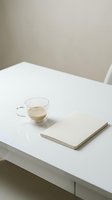 Minimalist workspace with glass cup of tea and blank notebook on white table against neutral beige wall, suitable for business and lifestyle design projects.