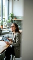 Happy young businesswoman laughing while using tablet in modern office cafe with plants and natural light for remote work and productivity.