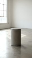 Minimalist concrete cylindrical pedestal in bright empty gallery space with natural light from large window, polished concrete floor, white walls, modern art display or product showcase setting.