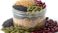 Glass jar filled with layered legumes and seeds including black sesame, quinoa, pumpkin seeds and red kidney beans on white background.