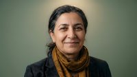Confident middle-aged South Asian woman smiling warmly, wearing a mustard scarf and gray blazer, portrait on a neutral green background.
