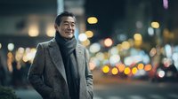 Smiling middle-aged Asian businessman in elegant gray coat and scarf walking on city street at night with warm bokeh lights creating atmospheric urban scene.