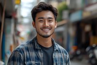 Young Asian man with bright genuine smile wearing plaid shirt and t-shirt outdoors in urban setting. Natural lighting highlights warm expression and modern casual style.