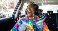 Happy African American woman laughing while listening to music with headphones in car, enjoying colorful tie-dye shirt and positive vibes during road trip.