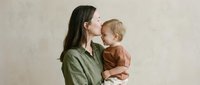 Young Caucasian mother in olive green shirt tenderly kissing blonde toddler child against neutral beige background in warm natural light studio portrait.