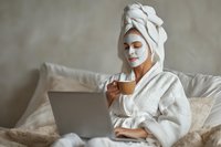 Young woman in bathrobe with facial mask relaxing in bed, drinking coffee and working on laptop during morning self-care routine.