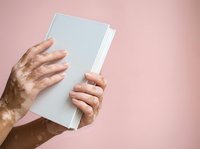 Person with vitiligo holding blank white book or magazine against pink background, showing skin condition diversity and inclusion in healthcare and medical education materials.
