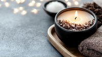Glowing aromatic candle in black ceramic holder decorated with coffee beans, spa towel and bath salt on gray background with bokeh lights.