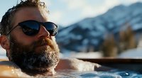 Bearded man relaxing in outdoor hot tub with steam rising, wearing sunglasses against snowy mountain backdrop during winter sunset. Wellness and luxury vacation concept.