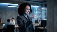 Woman with curly dark hair and medium skin tone wearing black blazer interacts with holographic data display showing financial charts and percentages in modern office environment with colleagues working at desks.
