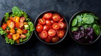 Fresh colorful salad ingredients in black bowls on dark background - cherry tomatoes, mixed lettuce leaves and herbs arranged in modern composition.