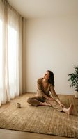 Young Asian woman in beige linen outfit sitting cross-legged on jute rug in minimalist living room with natural light streaming through sheer curtains.