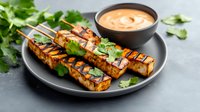 Grilled tofu skewers garnished with fresh cilantro served on black plate with creamy dipping sauce, healthy plant-based protein meal option.