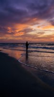 Silhouette of angler fishing at ocean shore during dramatic sunset with purple and orange clouds reflecting on wet sand and waves for outdoor recreation concepts.