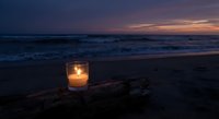 Glowing candle in glass holder on driftwood at twilight beach with purple sunset sky and ocean waves creating peaceful romantic atmosphere for meditation.