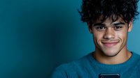 Young Latino man with curly dark hair and bright smile wearing blue sweater against turquoise background, looking at smartphone screen with positive expression.
