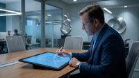 Mature male professional in dark business suit working on digital tablet with circuit board interface in modern office conference room with glass partitions and contemporary decor.