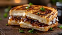 Grilled sandwich with caramelized onions and melted cheese on rustic wooden board, garnished with fresh herbs. Close up view showing golden-brown crust and gooey filling.