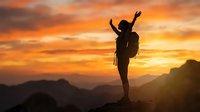 Silhouette of triumphant hiker with backpack celebrating success on mountain peak at golden sunset with dramatic orange sky and mountain ranges.