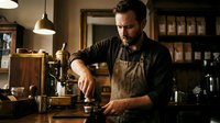 Professional male barista with beard preparing coffee in modern cafe interior with warm lighting and wooden counter.