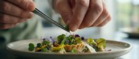Male chef with medium skin tone plating a gourmet salad with tweezers, adding microgreens and edible flowers to a fine dining dish. Professional culinary presentation.
