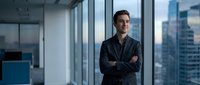 Confident young Asian businessman with crossed arms standing in modern office with city skyline view through large windows during daytime.