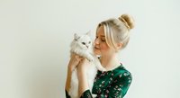 Young woman with blonde hair in a bun, fair complexion, holding a fluffy white cat against her shoulder. Studio portrait with neutral background, suitable for pet care, veterinary, adoption, and lifestyle content.