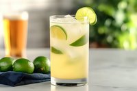 Refreshing lime cocktail with ice and citrus slices on kitchen counter. Fresh limes and beer glass in background create summer drink atmosphere for beverage marketing.