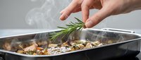 Hand adding fresh rosemary to roasted meat and vegetables in a baking pan with steam rising, suitable for culinary and food preparation content.