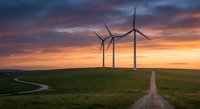 Three wind turbines stand on a green hill at dramatic sunset with vibrant orange sky, symbolizing clean renewable energy and sustainable power.