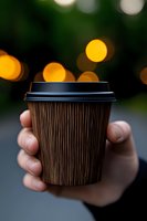 Hand holding brown rippled disposable coffee cup with black lid against blurred bokeh lights background, creating cozy autumn atmosphere.