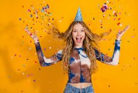 Excited young Caucasian woman with long blonde hair celebrating birthday party with confetti and party hat on bright orange background.