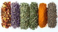 Colorful rows of dried herbs and spices including lavender, oregano, turmeric powder, and black pepper arranged on white background for culinary and seasoning use.