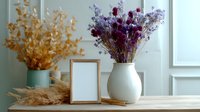 Minimalist home decor with dried purple flowers in white ceramic vase, golden wheat stalks and empty photo frame on wooden surface against soft blue wall.