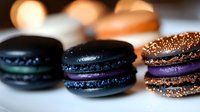 Elegant dark macarons with shimmering surfaces alongside glittery bronze treats on wooden surface with warm bokeh lights creating luxurious dessert display.