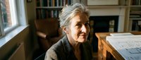 Mature woman with gray hair and light skin tone, wearing dark cardigan, sitting at wooden desk in home office with bookshelves and window, professional architect or designer reviewing blueprints.