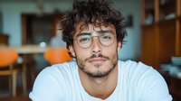 Young Mediterranean man with curly dark hair and glasses wearing white t-shirt looking directly at camera with confident smile in office environment.