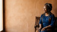 Middle-aged African American woman wearing denim shirt sitting on wooden chair against warm terracotta wall, looking away thoughtfully, suitable for lifestyle and portrait use.