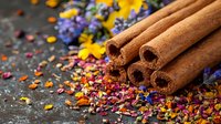 Aromatic cinnamon sticks with scattered colorful dried flower petals and spices on dark rustic background, macro food photography for cooking and aromatherapy.