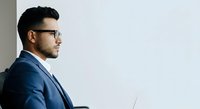 Young man with dark hair and beard, wearing glasses and navy blue blazer, sitting in profile against white background, suitable for corporate, business, and professional website design.