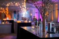 Elegant wedding reception venue with illuminated branches in tall vase, bokeh lights, and modern table settings creating romantic ambiance for special events.